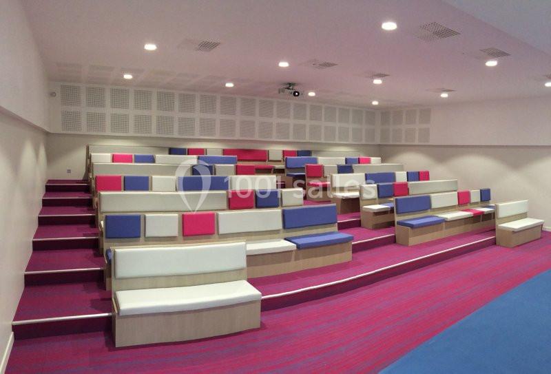 Salle de conférence moderne avec gradins colorés en rose, bleu et blanc, éclairage au plafond.
