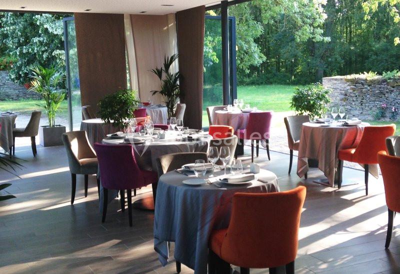 Salle de restaurant lumineuse avec des tables dressées, des chaises colorées et une vue sur un jardin verdoyant.