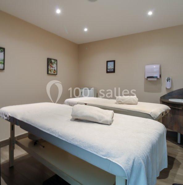 Deux tables de massage couvertes de draps blancs dans une salle lumineuse aux murs beige, décorée de cadres.
