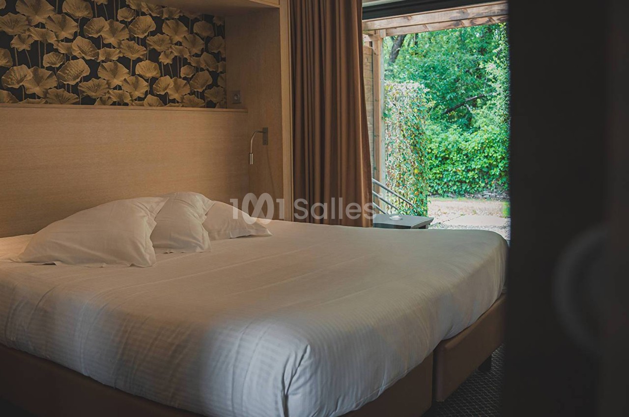 Lit double avec draps blancs dans une chambre au décor chaleureux, vue sur un jardin verdoyant par une porte ouverte.