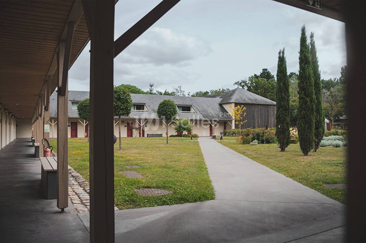 Allée en béton bordée de pelouses et d'arbres, menant à un bâtiment avec toit en ardoise et arcades en bois.