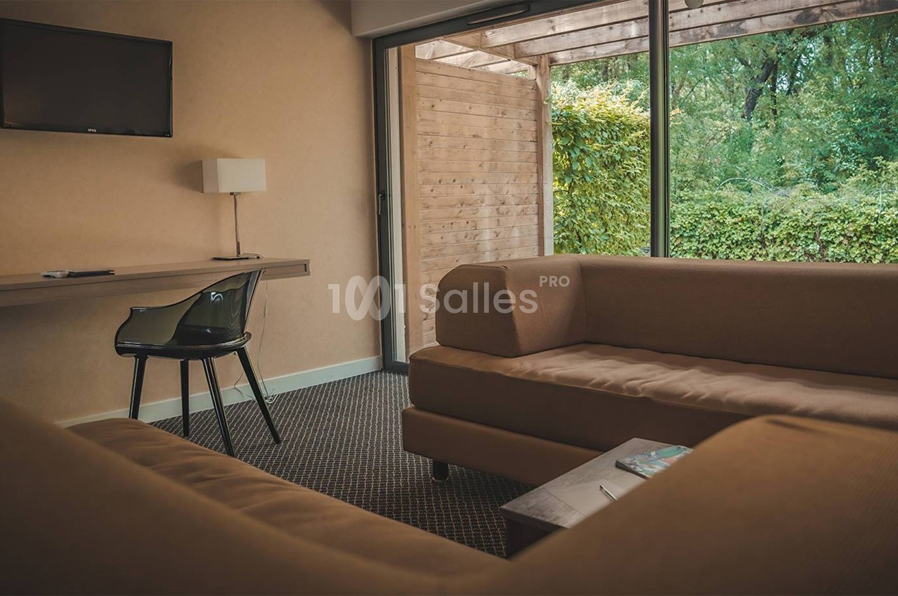 Salon d'hôtel avec canapés beiges, table basse, bureau, chaise noire et grande baie vitrée donnant sur un jardin.