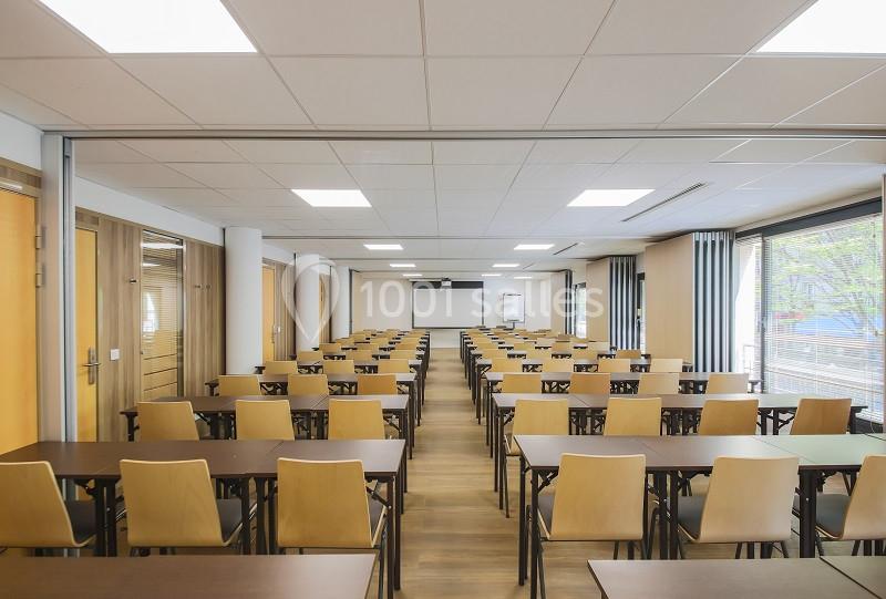Salle de conférence lumineuse avec des rangées de tables et chaises en bois, écran de projection au fond.