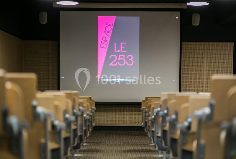 Salle de conférence vide avec des rangées de sièges et un écran affichant ’Espace Le 253’ en rose sur fond noir.