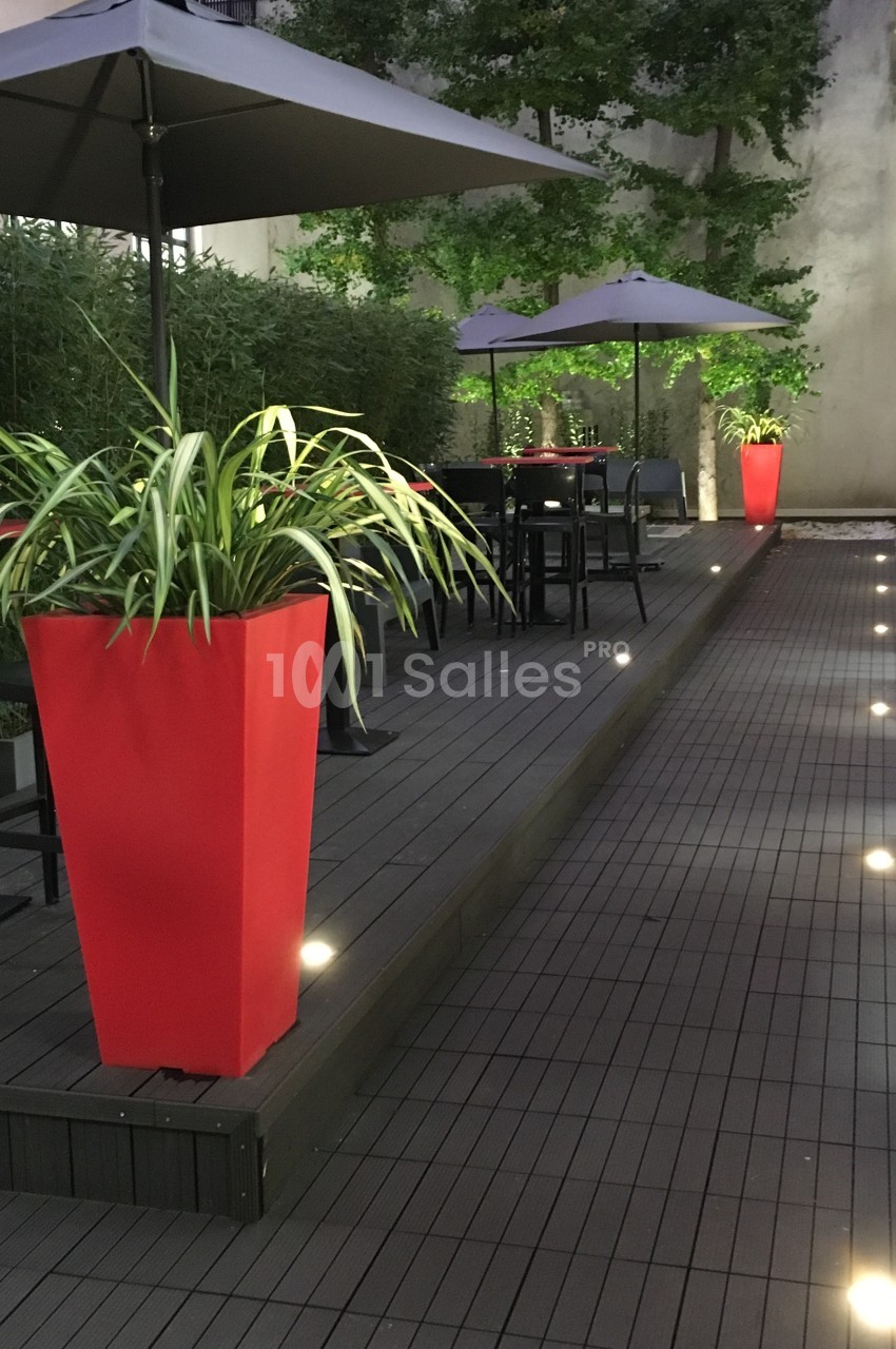 Terrasse en bois avec tables, chaises, parasols gris et plantes en pots rouges, éclairée par des lumières au sol.