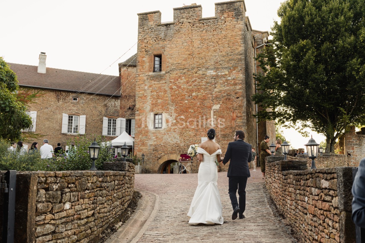 Un couple en tenue de mariage marche vers un bâtiment en pierre dans un cadre champêtre.
