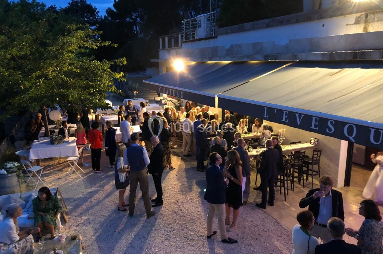 Groupe de personnes rassemblées en soirée près d'une terrasse éclairée, avec tables et arbres en arrière-plan.
