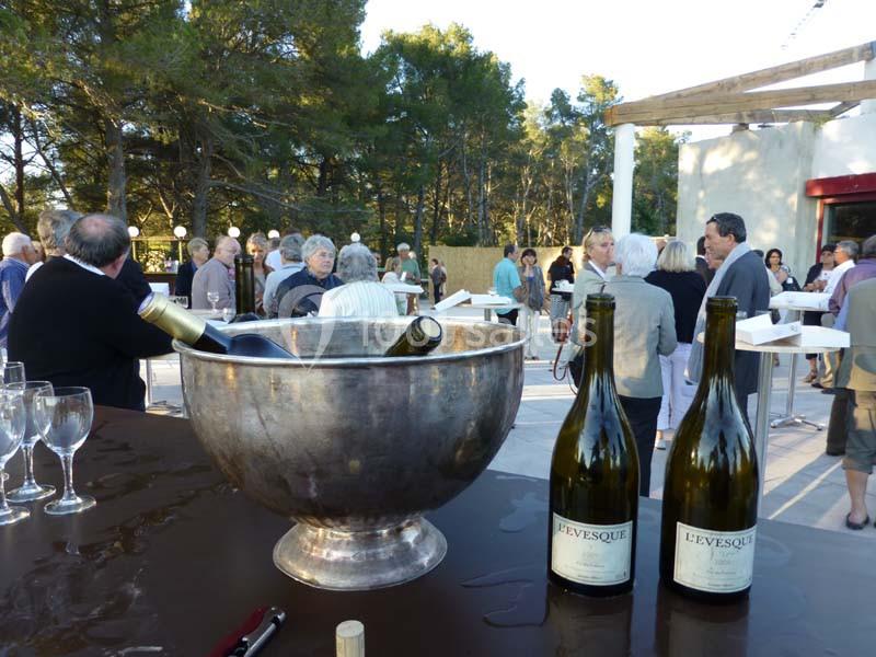 Un groupe de personnes discute en extérieur près de tables hautes, avec des bouteilles de vin et un seau à glace au premier…