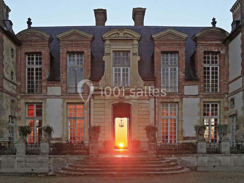 Façade d'un château ancien en pierre et briques, avec une lumière rougeoyante traversant l'entrée centrale.