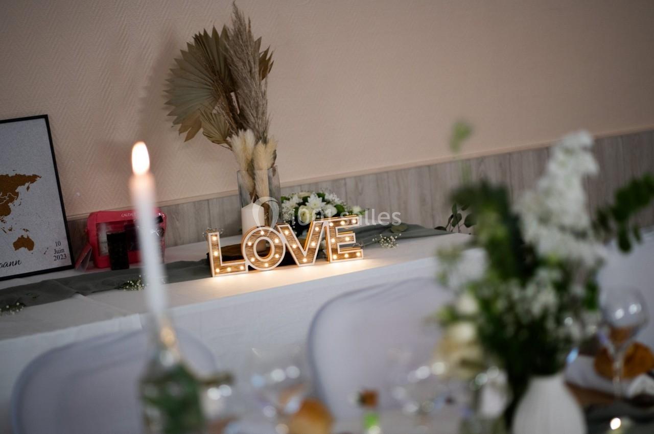 Décoration de table avec une enseigne lumineuse ’LOVE’, fleurs blanches et éléments naturels dans une salle de réception.