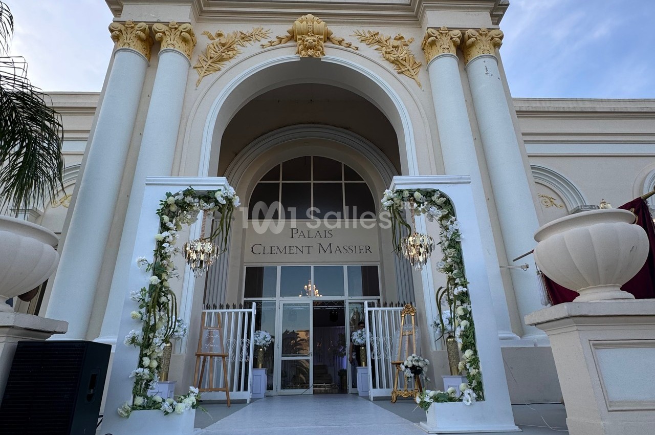 Entrée d'un bâtiment élégant avec colonnes blanches, décorations florales et enseigne ’Palais Clément Massier’.