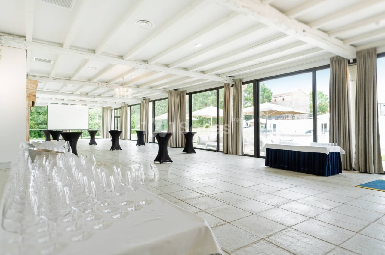 Salle lumineuse avec grandes baies vitrées, tables hautes noires, rangée de verres et vue sur une terrasse extérieure.