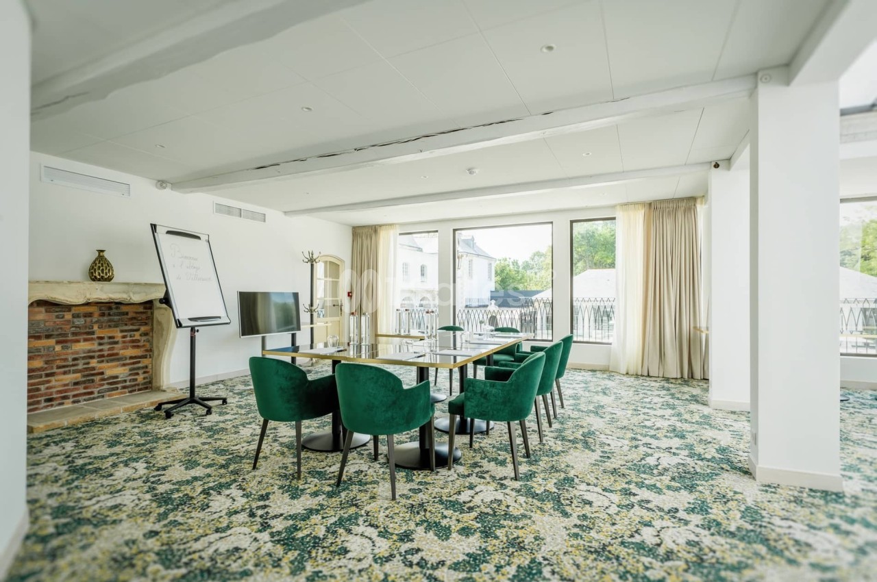 Salle de réunion lumineuse avec table, chaises vertes, tableau blanc, écran et grandes baies vitrées donnant sur l'extérieur.