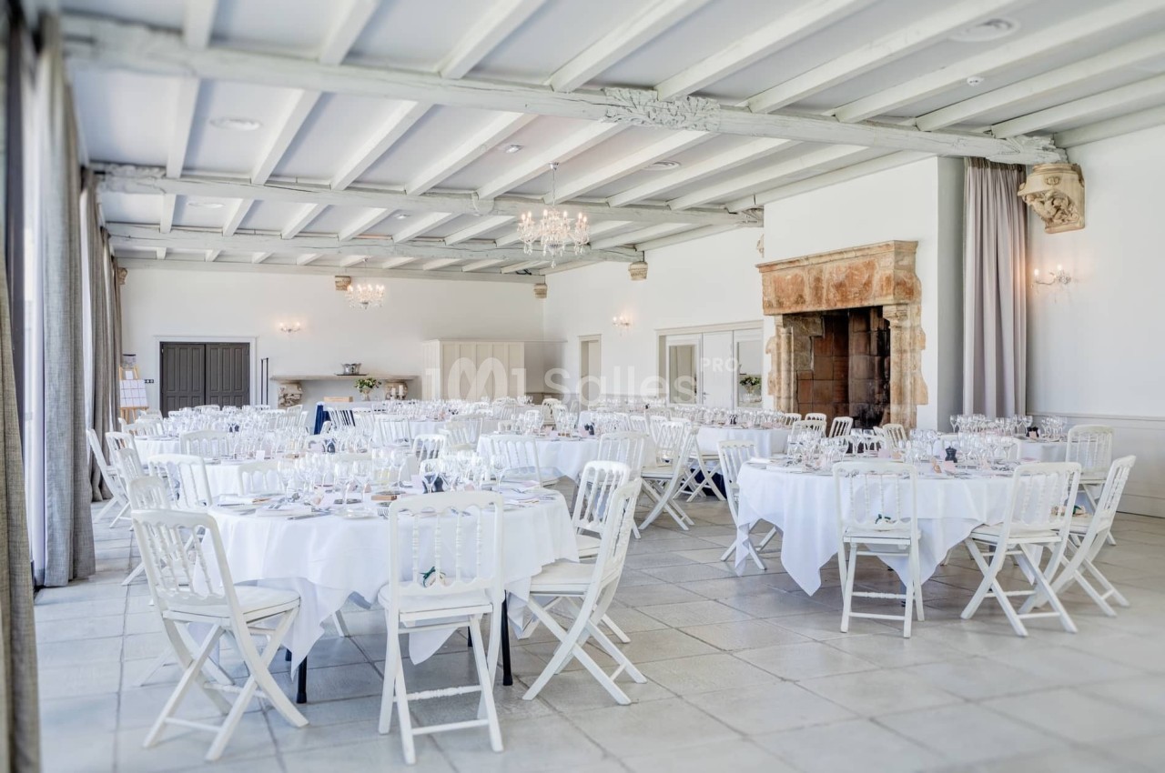 Salle de réception lumineuse avec tables rondes dressées, chaises blanches et cheminée en pierre.