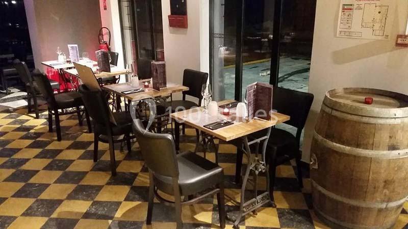 Salle de restaurant avec tables dressées, chaises noires, tonneau en bois et sol à carreaux jaunes et noirs.