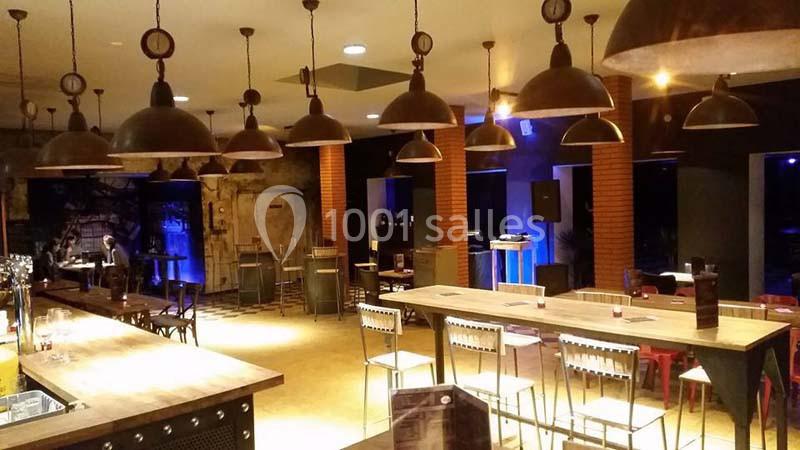 Salle de bar avec tables en bois, chaises métalliques et suspensions lumineuses dans une ambiance tamisée.