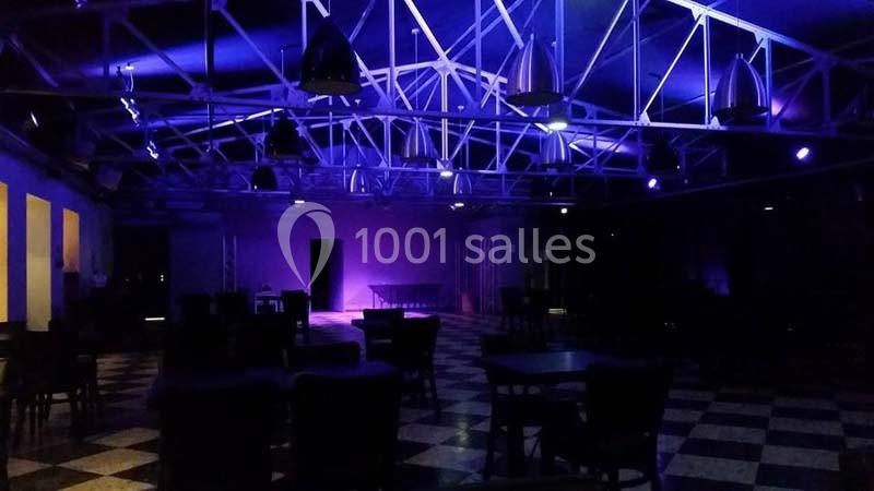 Salle sombre avec éclairage violet, plafond métallique, tables et chaises dispersées sur un sol à carreaux.