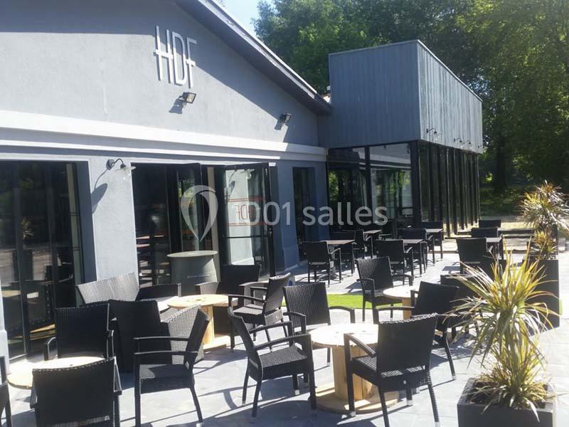 Terrasse extérieure d'un restaurant moderne avec des tables et chaises en rotin noir, entourée de végétation.