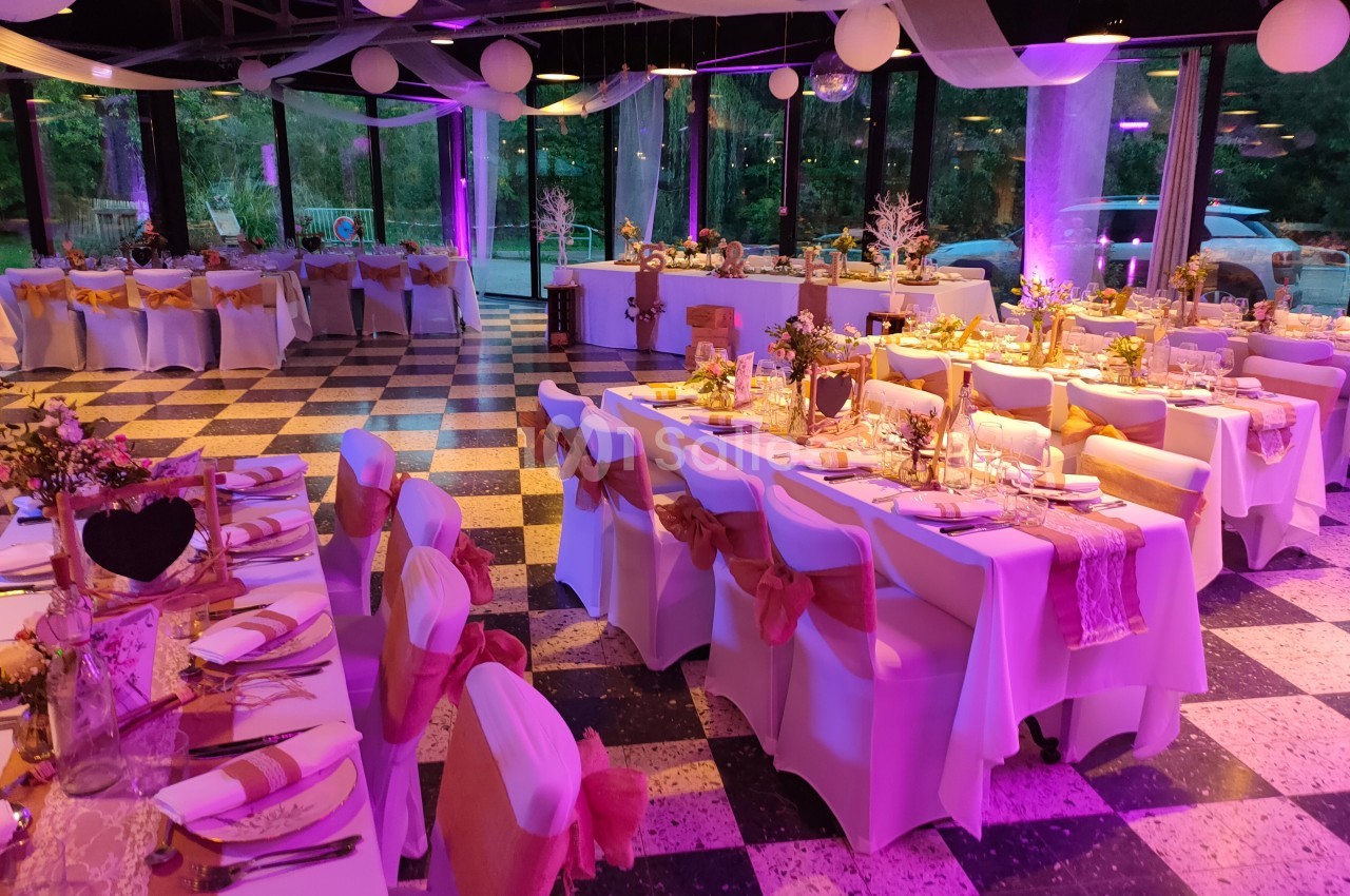 Salle de réception décorée pour un événement, avec tables dressées, chaises ornées de rubans et éclairage rose.