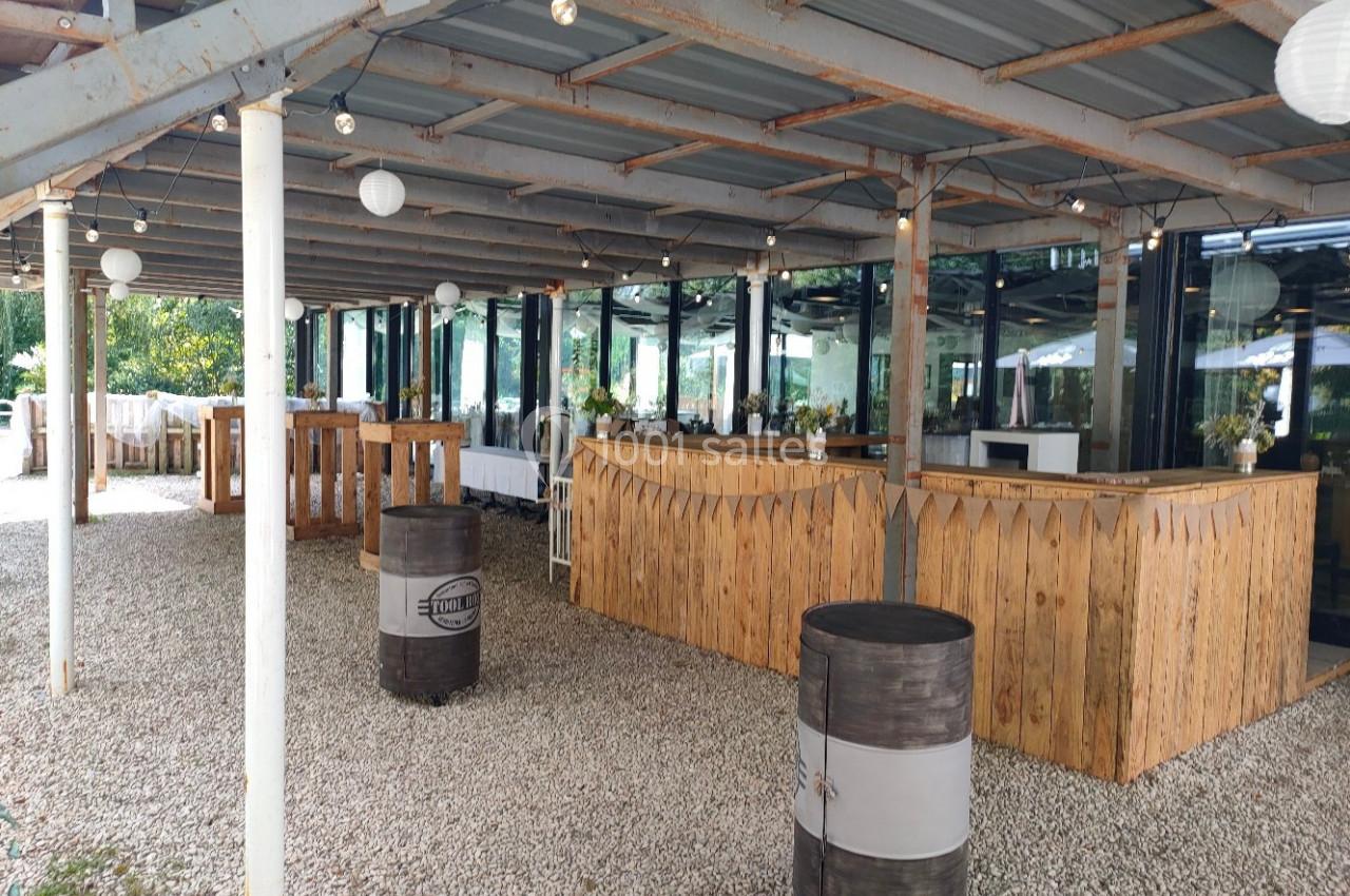 Terrasse couverte avec bar en bois, tables hautes et décorations lumineuses, sol en gravier.