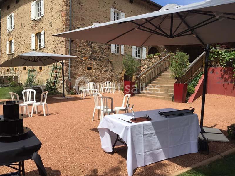Cour extérieure avec tables et chaises sous des parasols, devant un bâtiment en pierre avec un escalier.