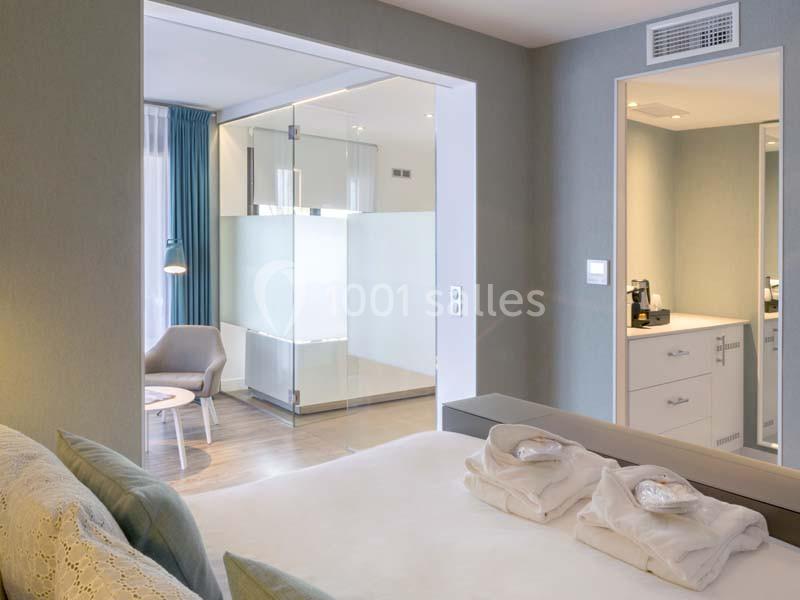 Chambre d'hôtel moderne avec lit double, salle de bain vitrée, fauteuil et coin café.