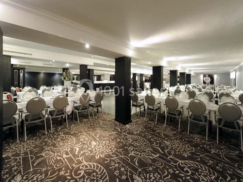 Salle de réception spacieuse avec tables rondes dressées, chaises grises et décoration élégante sur un tapis à motifs.