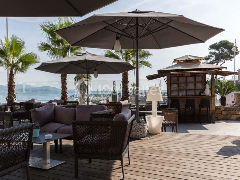 Terrasse en bois avec mobilier de jardin, parasols, bar extérieur et vue sur la mer bordée de palmiers.