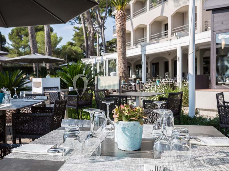 Terrasse d'un restaurant avec tables dressées, entourée de végétation et d'un bâtiment moderne en arrière-plan.