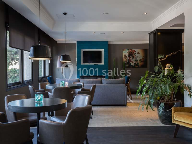 Salle moderne avec tables rondes, chaises en cuir, canapé, télévision murale et plantes décoratives.