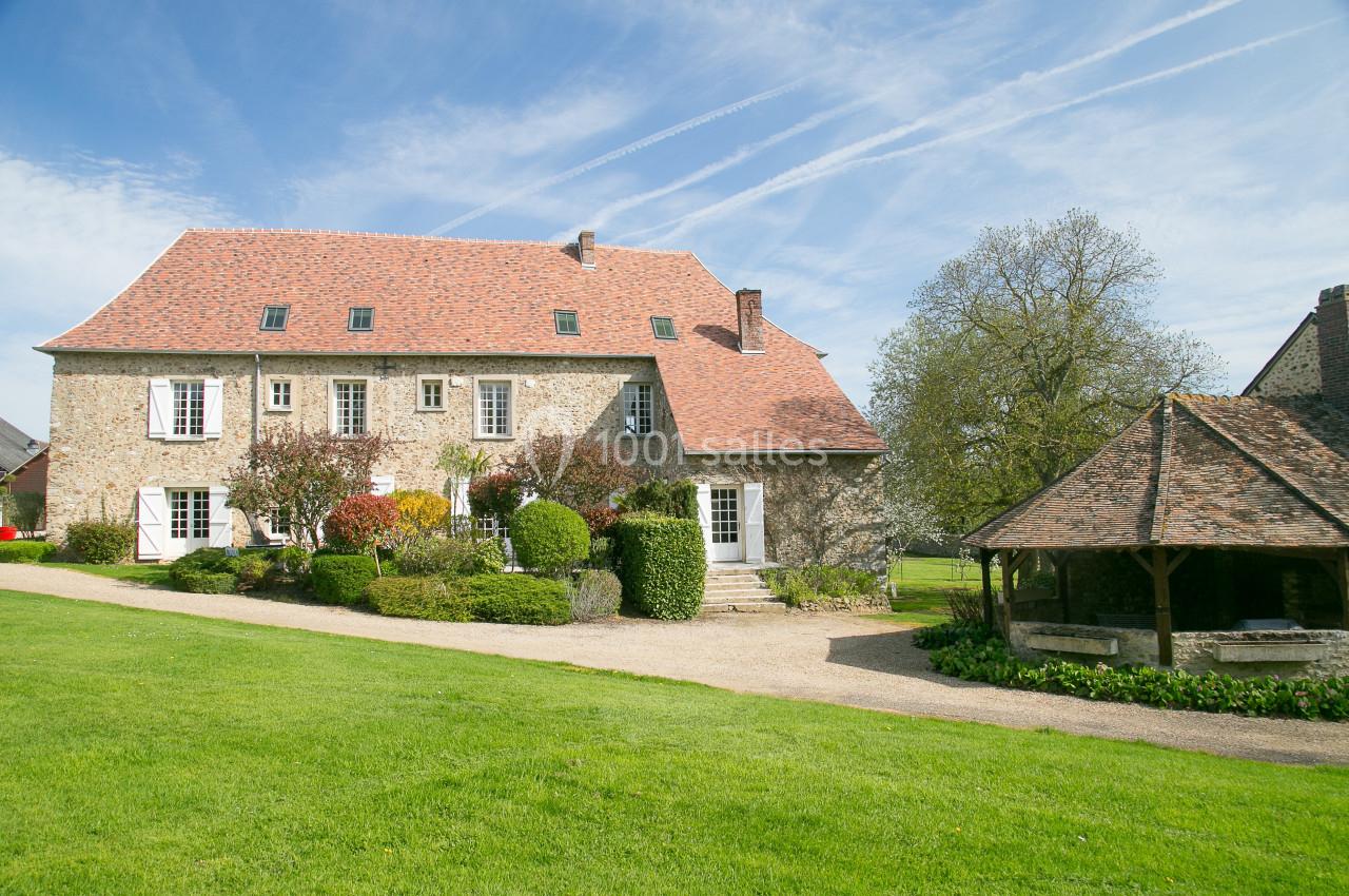 Grande maison en pierre avec toit en tuiles rouges, entourée de verdure et d'un puits couvert dans un jardin.