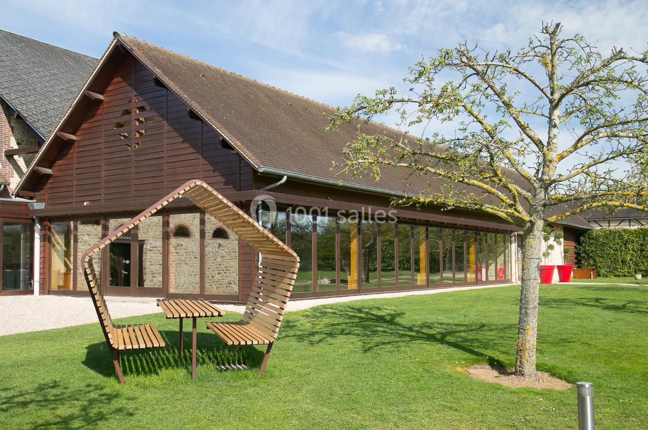 Bâtiment en briques et bois avec de grandes baies vitrées, devant un jardin aménagé avec un banc design et un arbre.