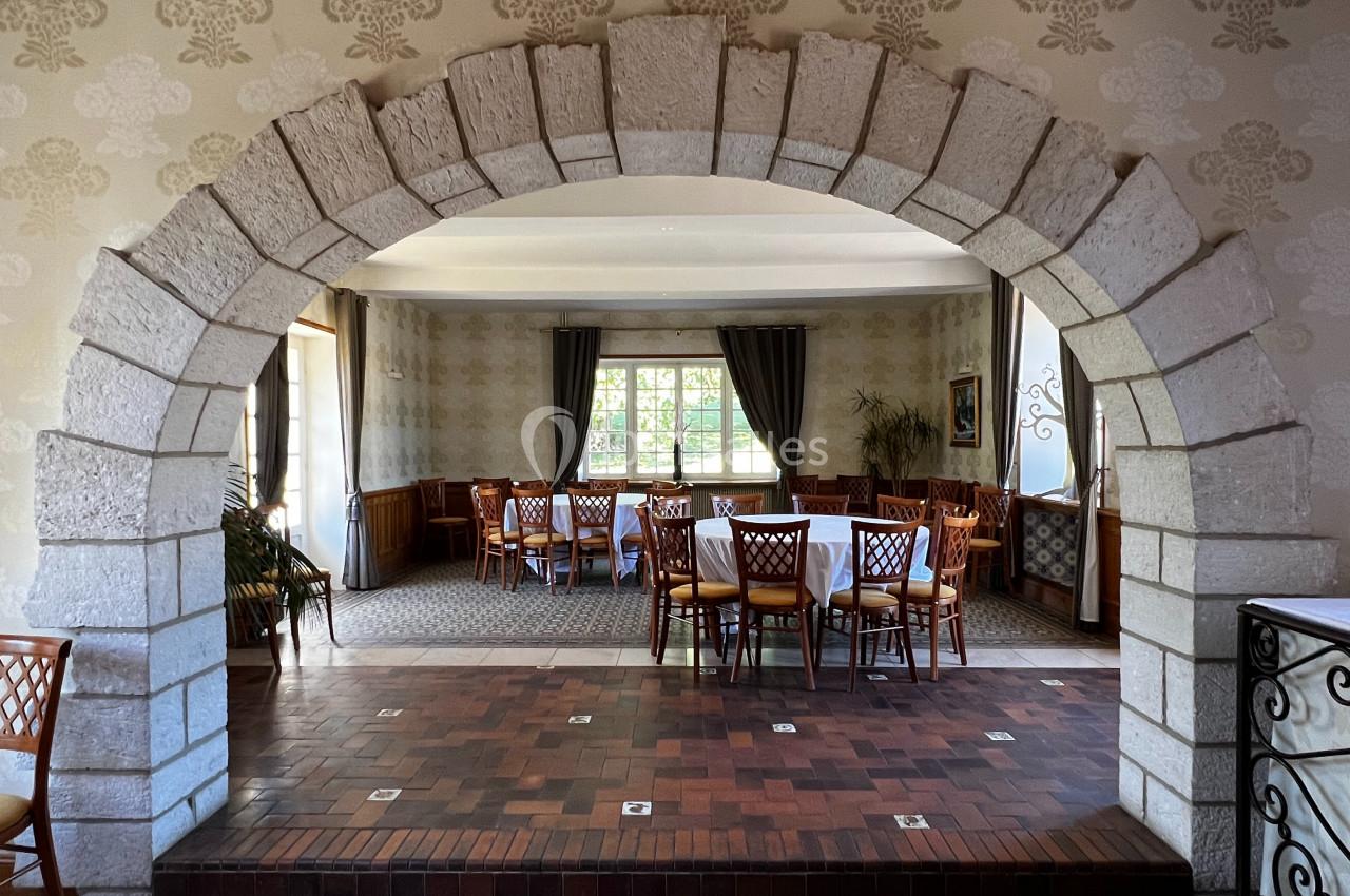 Salle à manger lumineuse avec des tables et chaises en bois, vue à travers une arche en pierre.