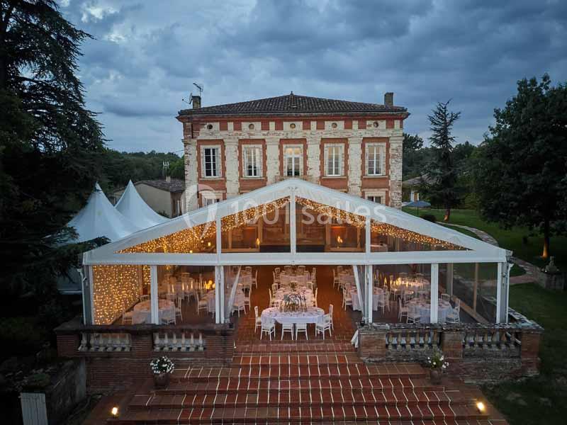 Vue d'une grande tente transparente illuminée installée devant une maison en briques, entourée de verdure.