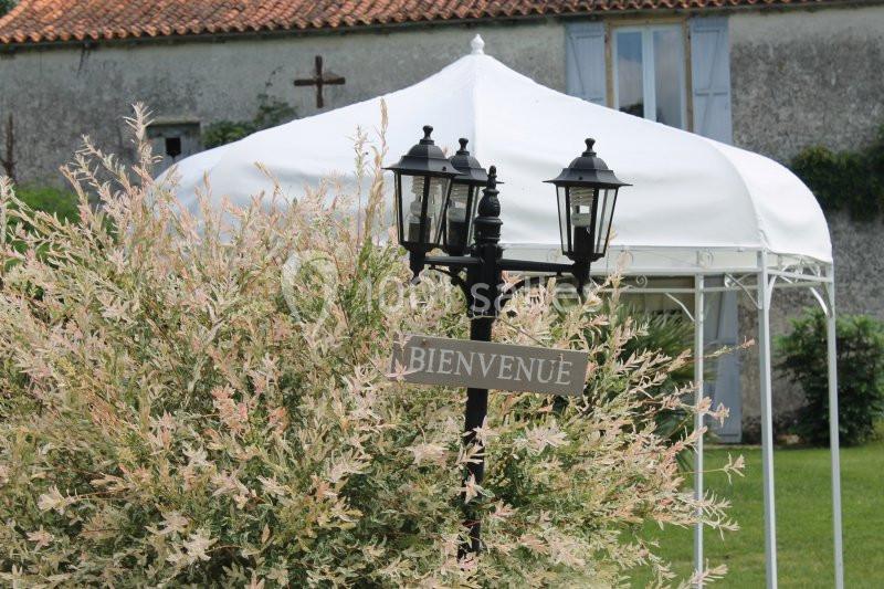 Lampadaire noir avec un panneau ’Bienvenue’ devant un arbuste et une tonnelle blanche dans un jardin verdoyant.