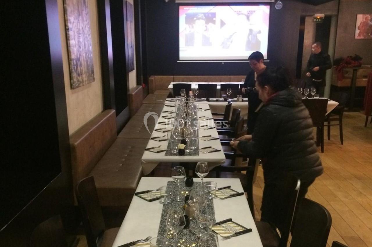 Salle de restaurant avec une longue table dressée, deux personnes ajustant les couverts et un écran en arrière-plan.