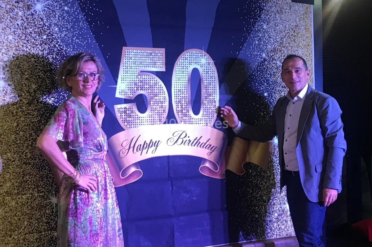 Un homme et une femme posent devant une décoration ’50 Happy Birthday’ avec des paillettes dorées en arrière-plan.