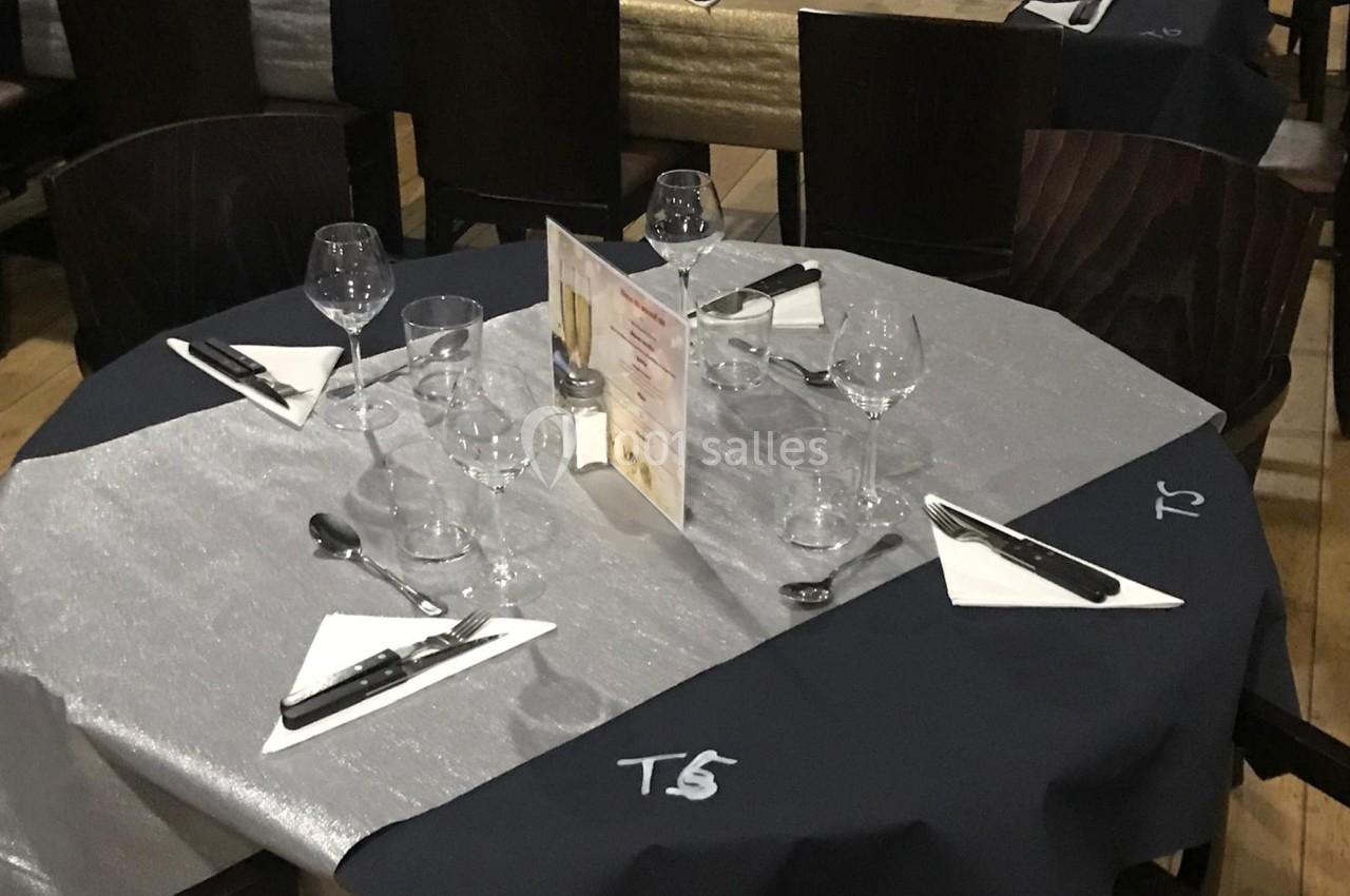 Table de restaurant dressée avec nappes noires et grises, verres, couverts, serviettes pliées et menu au centre.