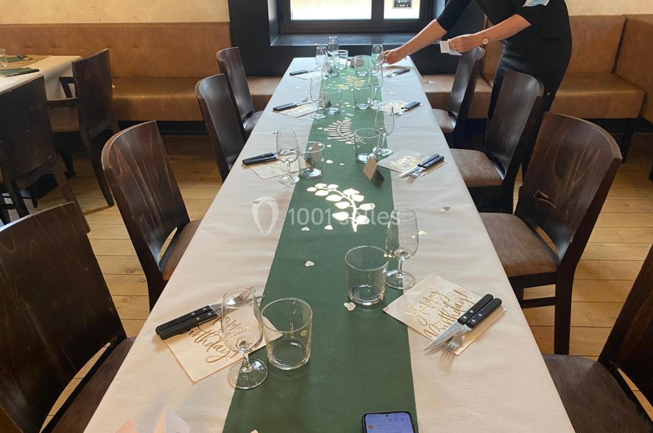 Table décorée pour un repas, avec chemin de table vert, vaisselle, menus et une personne ajustant les détails.