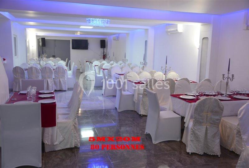 Salle de réception décorée en blanc avec des tables dressées, nappes rouges et chandeliers, capacité de 80 personnes.