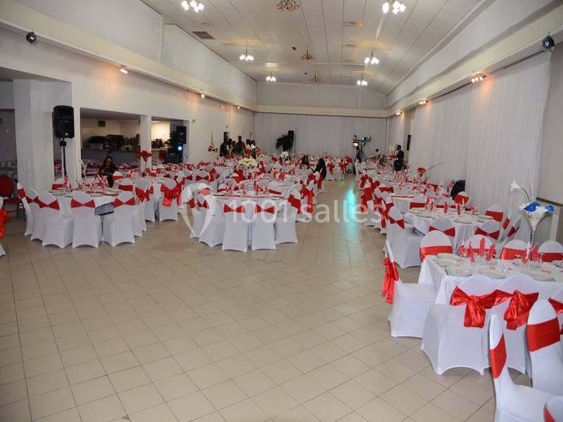 Salle de réception décorée avec des nappes blanches et des nœuds rouges sur les chaises, tables dressées pour un événement.