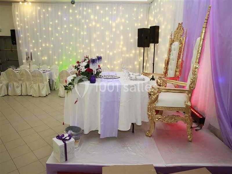 Salle de réception décorée avec une table ronde ornée de fleurs et un grand fauteuil doré sur une estrade.