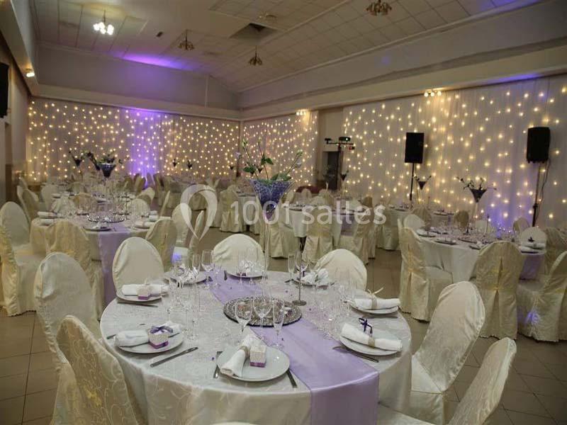 Salle de réception décorée avec des tables rondes, nappes blanches, chaises habillées et guirlandes lumineuses au mur.