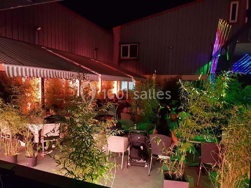 Terrasse extérieure éclairée la nuit, entourée de bambous, avec des tables occupées sous des auvents rayés.