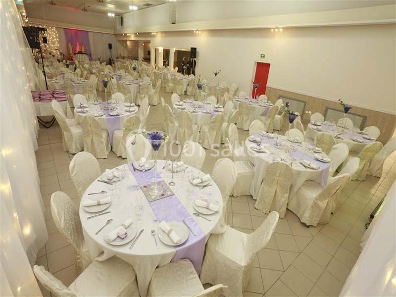 Salle de réception décorée avec des tables rondes, nappes blanches, chemins de table violets et chaises recouvertes.