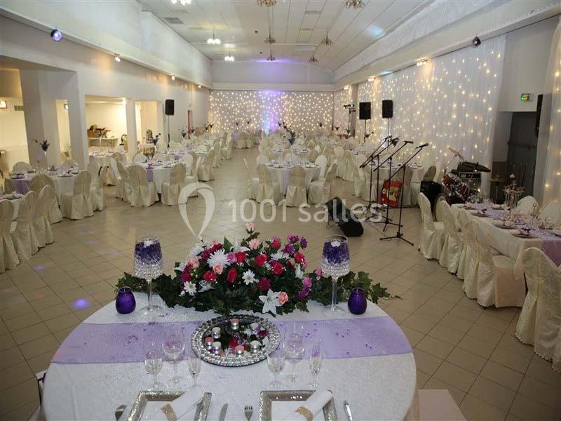 Salle de réception décorée avec des tables rondes, nappes blanches, fleurs et guirlandes lumineuses en arrière-plan.