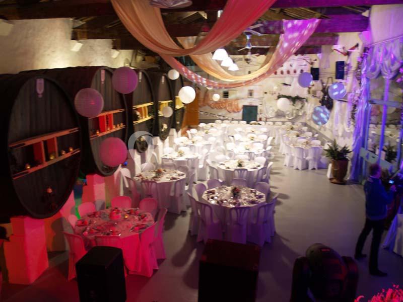 Salle de réception décorée avec des tables rondes dressées, des tonneaux en bois et des guirlandes lumineuses suspendues.