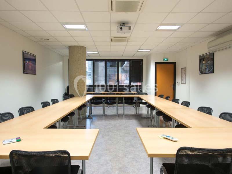 Salle de réunion lumineuse avec tables disposées en U, chaises noires, tableau blanc et projecteur au plafond.