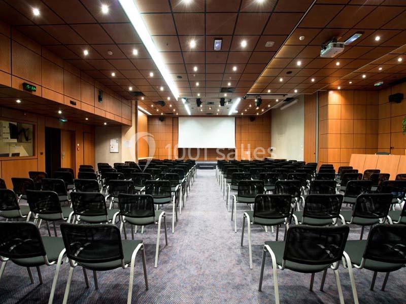Salle de conférence vide avec rangées de chaises noires, écran de projection et éclairage au plafond.