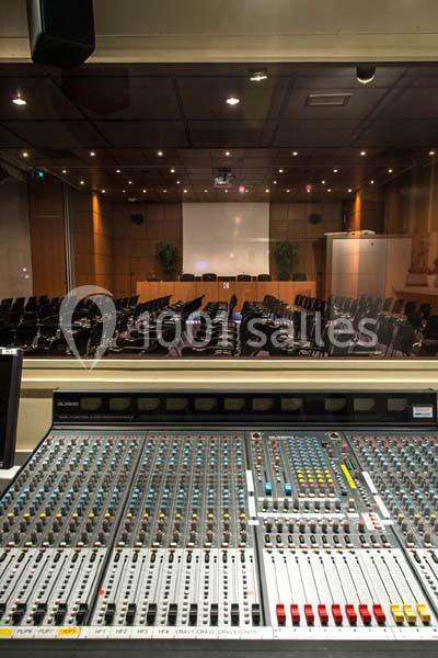 Vue d'une régie audio avec une console de mixage en premier plan et une salle de conférence vide en arrière-plan.